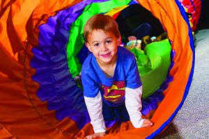 Boy wearing superman shirt crawling through a tunnel | Pediatric Therapy Center’s occupational therapy program includes sensory integration therapy to help a child further develop sensory motor skills.
